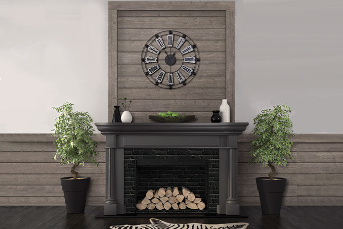 A modern looking fireplace with a dark gray mantel. There is a shiplap border behind the mantel with a metal clock hanging from the wall. Two small green trees frame the fireplace and a zebra stripped rug is on the ground as an accent piece. 