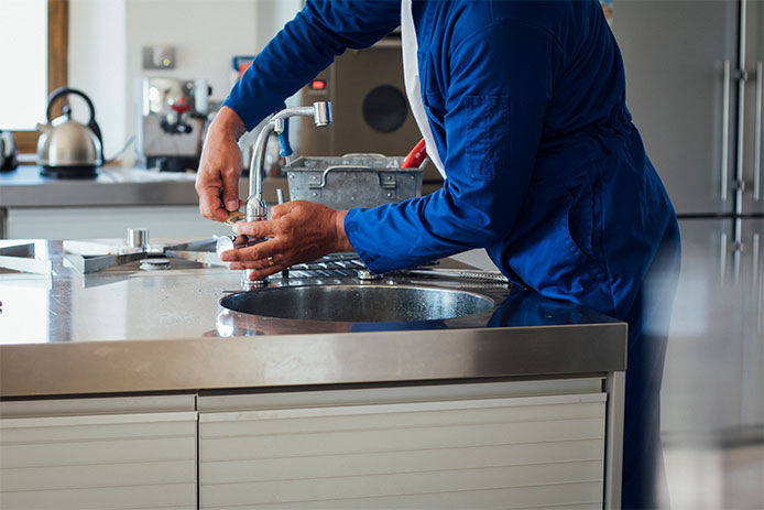 A shot of a man's hands working on a kitchen sink. He is wearing blue overalls with his tool kit in the background.