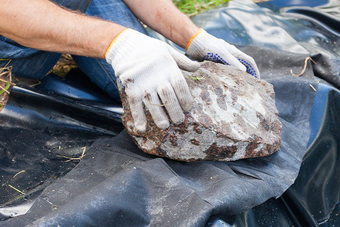 a close-up of a man's hands holding a large rock over a pond liner, pressing it down to hold the liner in place. The focus is on the rock and liner, with no visible background. The man's hands are visible, wearing white gloves as he carefully places the rock over the liner. The liner is neatly arranged and tucked in, indicating that the man has taken great care in the installation of the pond.