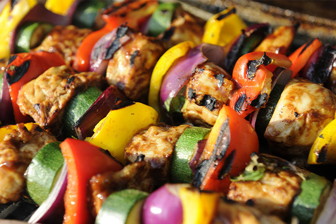 Colorful kabob skewers are shown cooking atop a grill. Dark grill marks on the meat and vegetables include yellow squash, red pepper, green zucchini, and purple onion. 