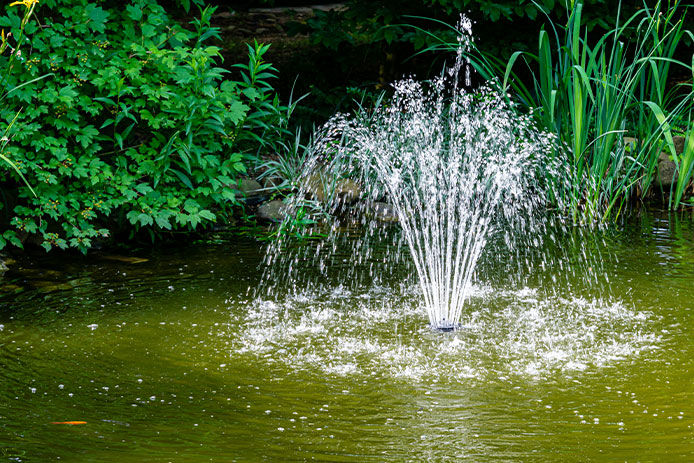 a beautiful sprinkler fountain in the middle of a pond, surrounded by lush green foliage that gives the water a green tint. The fountain creates a mesmerizing display of water droplets shooting up into the air and falling back into the pond, creating ripples that further enhance the beauty of the pond. 