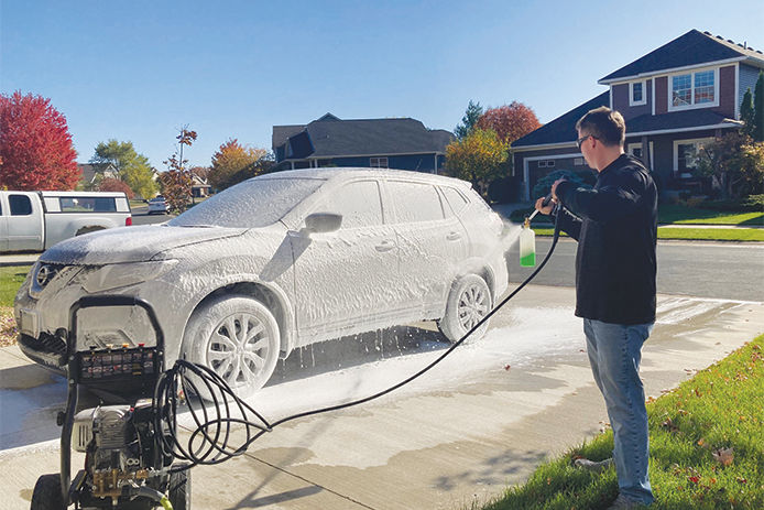 Man using a gas pressure washer to wash his car on a hot sunny day. The car is covered in soap and the driveway is wet.