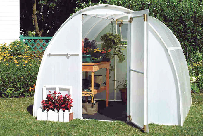 A white pvc and canvas greenhouse with the door open and a small wooden table filled with plants and garden tools on the inside