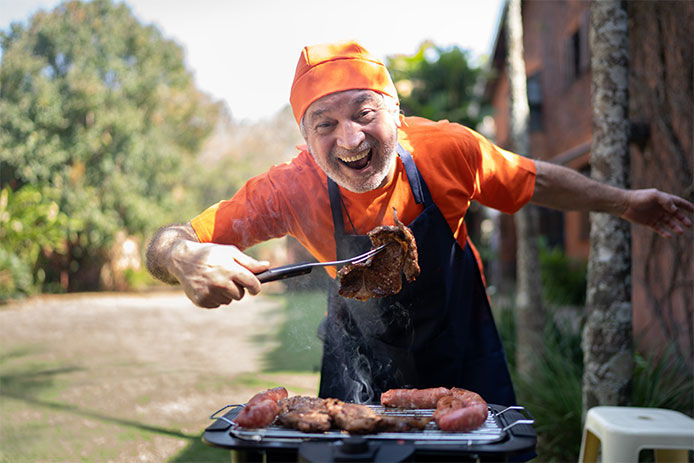 Man smiling, wearing an apron and holding a piece of meat on a grill fork