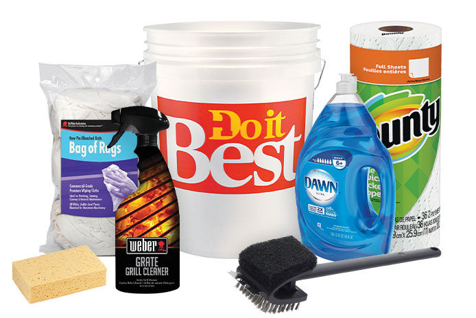 An assortment of grill-cleaning supplies is shown against a white background, including a yellow sponge, a bag of white rags, a brush, and a white Do It Best bucket. The supplies include Bounty paper towels, Dawn liquid soap, and Weber grill cleaning spray in a black spray bottle.