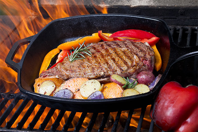 Cast iron skillet cooking a pork tenderloin with pepers, potatoes, and rosemary over a charcoal stove