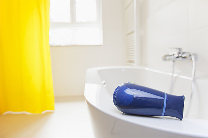 A blow dryer lying on the edge of a bathtub in a all white bathroom