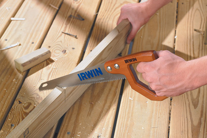 A man using a Irwin hand saw to cut a 2x4 piece of lumber on a wooden deck 