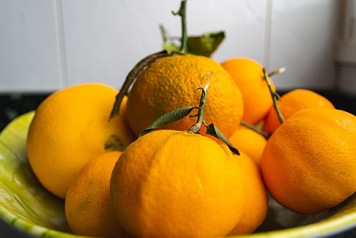 A yellow bowl is filled with bright orange clementines. Some of the fruits still have leaves and stems attached. The bowl sits in a pantry with a white shiplap wall.