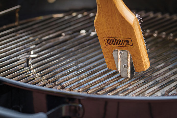 A wire and wooden grill brush being used to clean and season a black charcoal grill