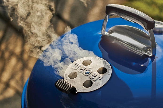 A close up image of a blue Weber charcoal grill lid with smoke coming out of the air release valve