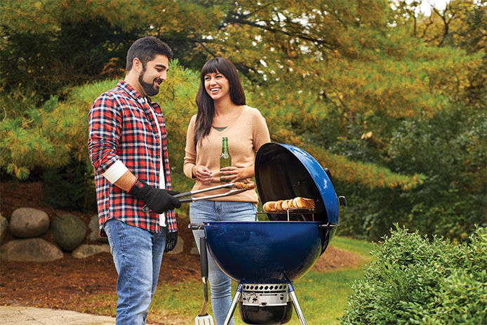 A happy couple standing around a blue charcoal grill grilling hot dogs and drinking beer on a hot summer day 