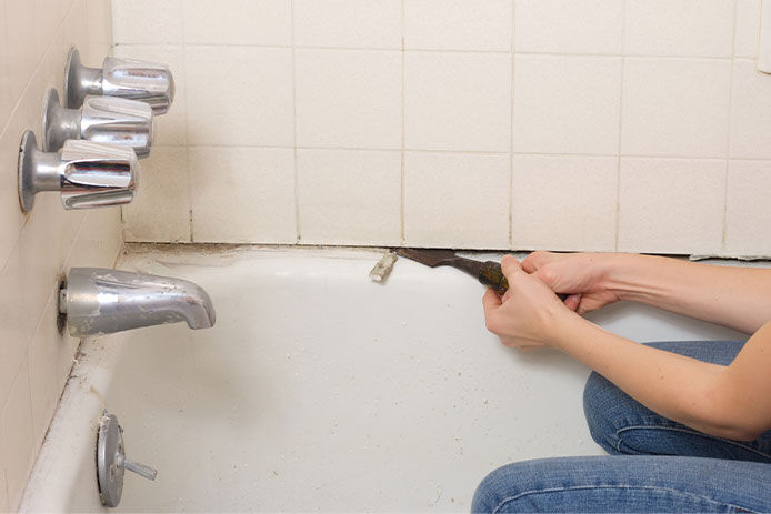 Young woman working to fix the bathtub caulk with toxic mold with a chisel and is involved in hard work and dirt