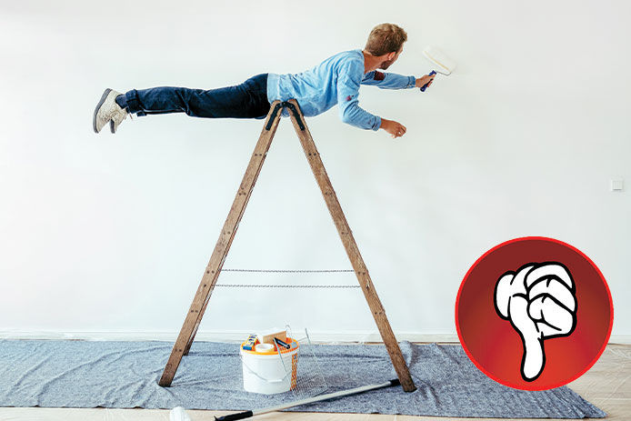 A man wearing a blue shirt and jeans incorrectly laying across the top of a step ladder painting the wall