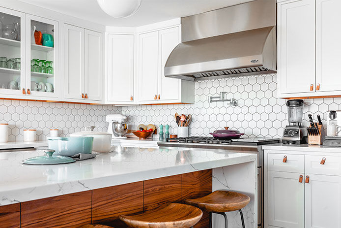 A clean, white kitchen is shown with white cupboards and a marble island featuring wooden barstools. The counters are pristine.