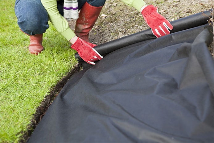 Person wearing red gardening gloves crouched laying down landscaping fabric