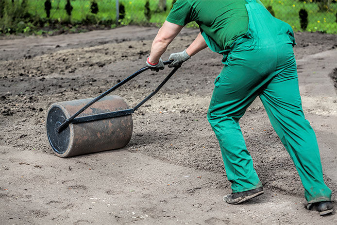 Person wearing green overalls using a yard roller to flatten the soil