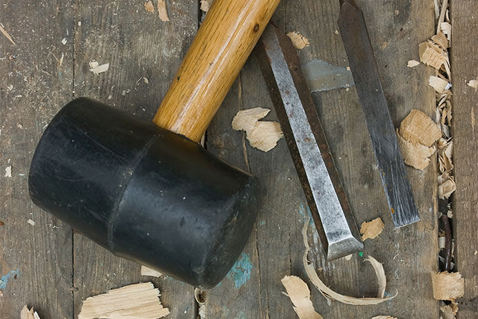 A handheld mallet sitting on a wooden table next to two wood chisels and wood shavings all around