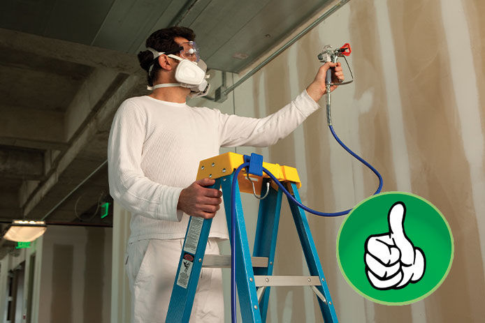 A professional painter wearing a N95 mask standing on a blue and yellow ladder correctly holding onto the ladder while painting a wall with a paint sprayer