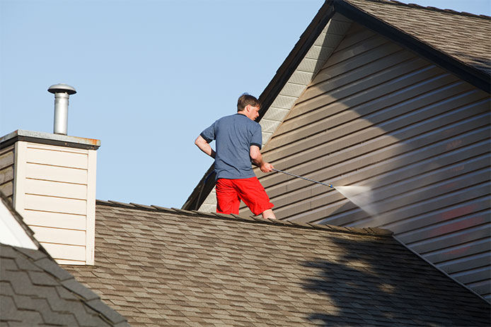 A middle age man wearing a blue shirt and red shorts standing on top of his roof using a power washer to clean the siding of his home