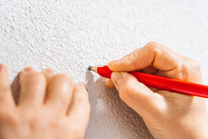 A person’s hands are holding a red carpenter pencil, marking white stucco. 