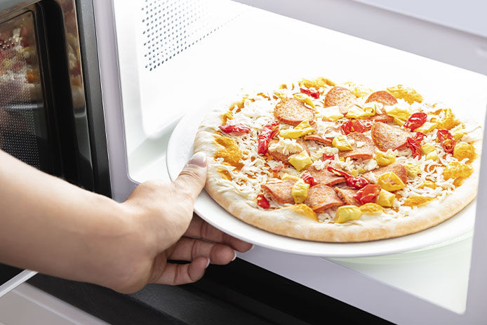 A person placing a microwaveable frozen pizza into the bright inside of a microwave. The round pizza has mixed meat and cheese toppings. 