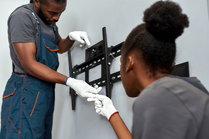 Close view African man and woman during TV wall brackets installation work. Female assistant giving tools to handyman, both in white protective gloves. Team work concept.