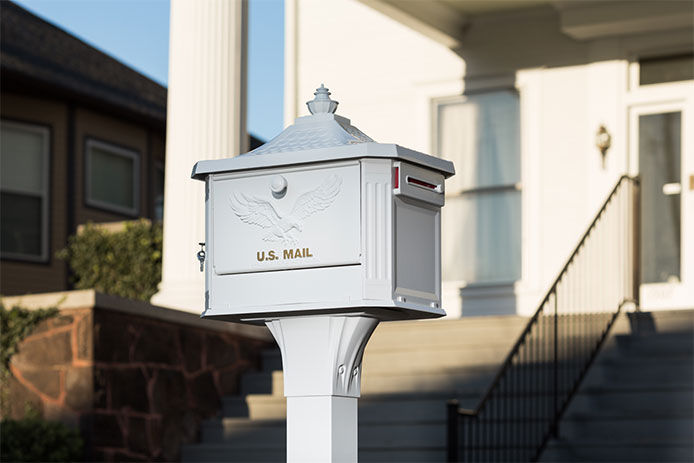 A white mailbox stands in front of a house. The locking mailbox is mounted atop a post with an eagle on the front.