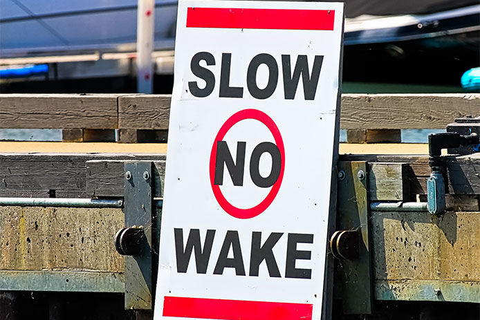 A slow no wake sign on a dock with boats in the background.