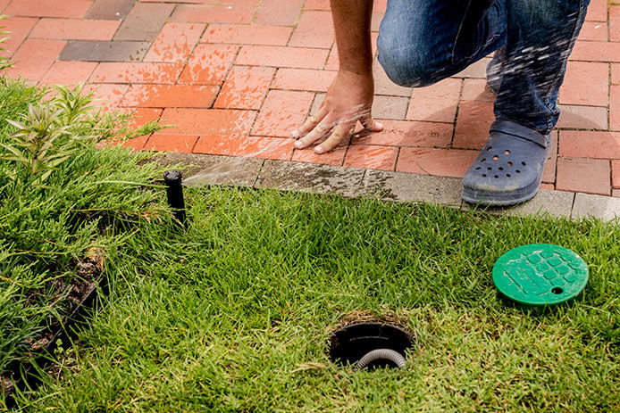 A man crouched down next to an in ground sprinkler head with the lid to the water shut off open. He is draining water out of the system for the winter time.