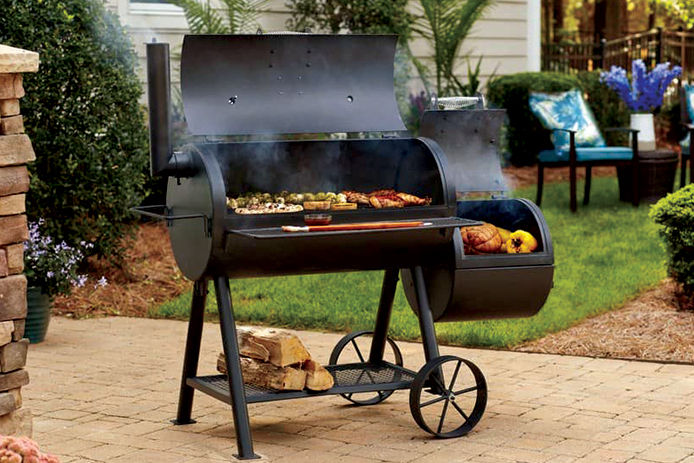 A black duel house smoker on a back patio with meats and vegetables on the grates. 