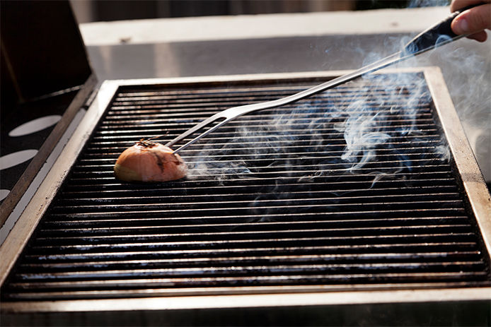 A person holds a long-handled fork attached to half an onion. They are rubbing the onion along a grill's grates as smoke rises from the hot surface.