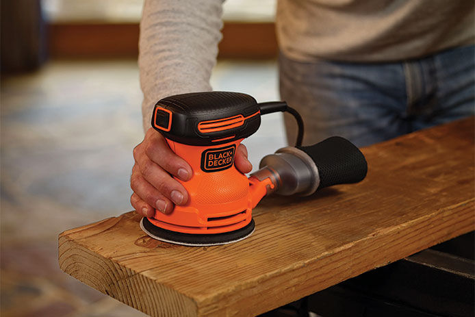 A homeowner useing a corded black and decker orbital sander with dust collection on a piece of stained wood 