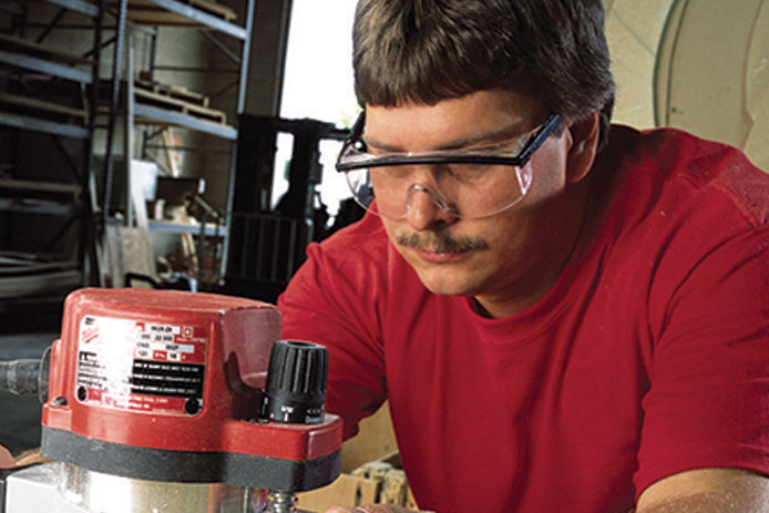 A man in a red shirt with safety goggles uses a red and silver wood router. 