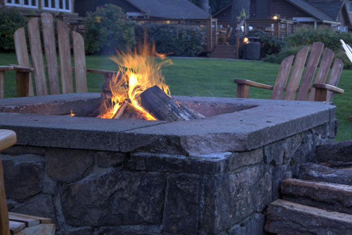 Two Adirondack chairs are shown at dusk near a large grey above-ground fire pit. There is a fire burning in the pit with a pleasant orange flame.