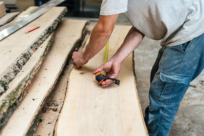 A man wearing a t-shirt and jeans using a tape measure to measure out a slab of live edge wood 