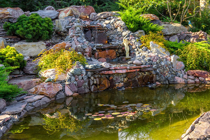 showcases a stunning backyard rock pond, featuring three layers of cascading rocks and water plants surrounding the area. The pond is adorned with a variety of rocks, both small and large, creating a natural look and feel. The water in the pond is clear and still, with a reflection of the lush greenery in the surrounding area. The rocks around the pond provide texture and depth, creating a visually appealing landscape.