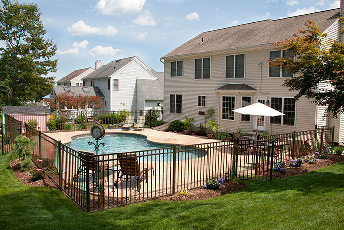 Lush, resort-like backyard salt water swimming pool with flagstone stamped concrete patio behind colonial style home. Bronze fencing is seen around the pool area of this suburban backyard.