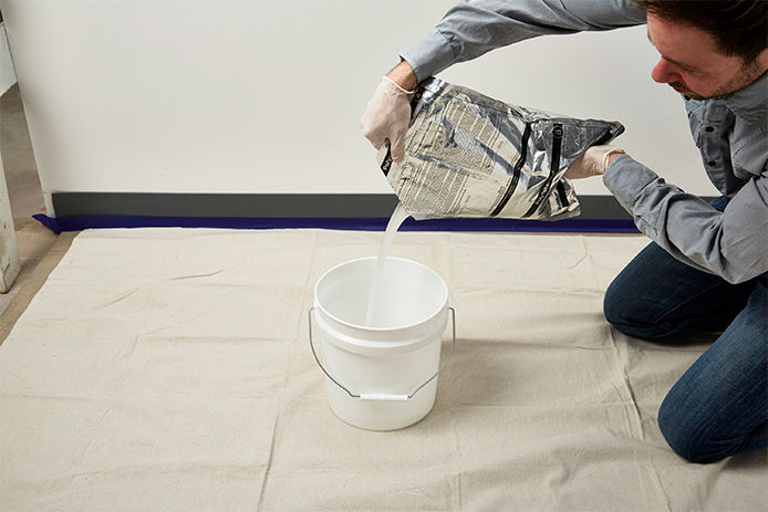 A man pouring the contents of a floor coating package into a one gallon white bucket on the floor of a garage