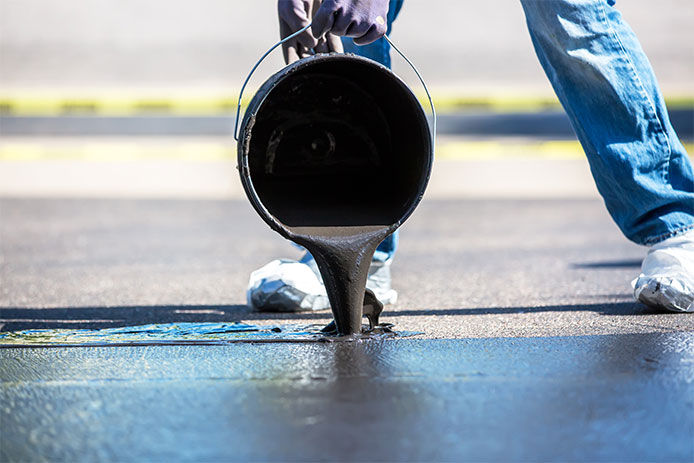 A person wearing jeans and white sneakers is pouring a bucket of black asphalt resurfacer onto the pavement. The thick resurfacing material covers the blacktop, creating a smooth, shiny surface.