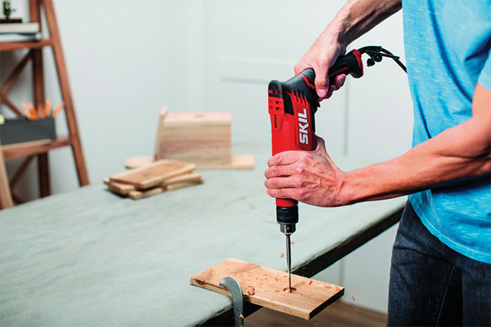 A man using a corded SKIL drill to drill a hole through a piece of wood that is clamped down to a work table