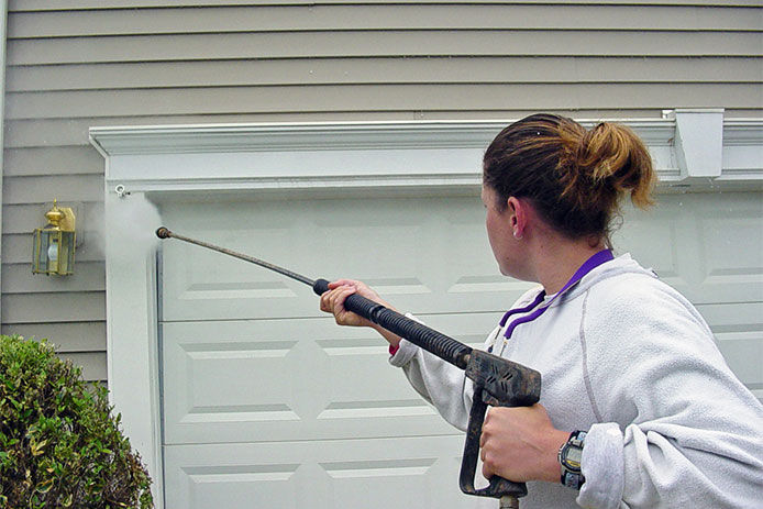 A middle aged woman using a pressure washer to wash down the siding around the garage