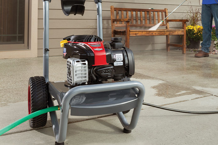 A gray, red and black gas pressure washer on a residential driveway attached to a green garden hose. The owner is pressure washing a wooden bench in the background.