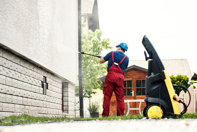 A professional house cleaning wearing red coveralls and power washing a brick house with a black and yellow power washer.