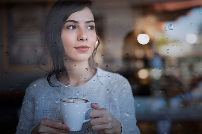 Young Woman standing in front of window and Enjoying A Cup Of coffee At Home on a rainy day