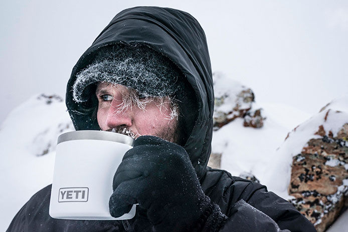 Man bundled in winter gear drinking from a YETI mug with a cold and icy background