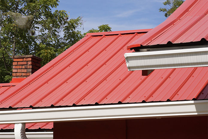 A close up image of a red metal roof top with white gutters and a brick fireplace with a blue sky and green tree in the background