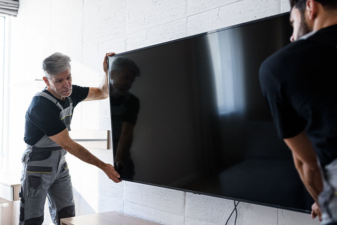 Two professional technicians, workers in uniform installing television on the wall indoors. Construction, maintenance and delivery concept