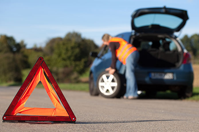 Orange caution triangle on the side of the road with a person in the background changing their tire blurred