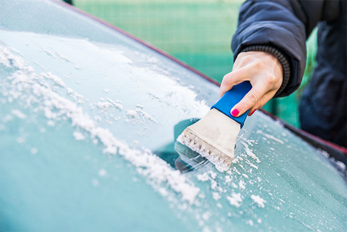 Frost built up on a cars window shield.  A woman is using an ice scraper to remove the frost.